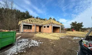 Budowa domu z cegły, widoczny szkielet dachu, drewno budowlane, kontener na śmieci.