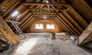 Poddasze 100m2 do adaptacji. Widok na drewnianą konstrukcję dachu, okna, drabiny i gruzy. Koszt adaptacji poddasza 100m2 może być wysoki.