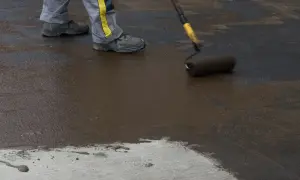 Pracownik maluje podłogę w piwnicy na gruncie ciemną masą. Widać jego nogi w roboczych spodniach i buty.