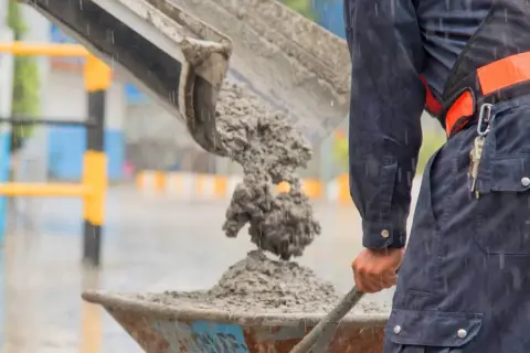 Robotnik w deszczu wylewa świeży beton do taczki. To właśnie jest beton – mieszanka cementu, kruszywa i wody.