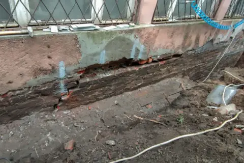 Widoczny pęknięty fundament z cegły, odsłonięty po wykopaniu ziemi. Widać ślady napraw i narzędzia.