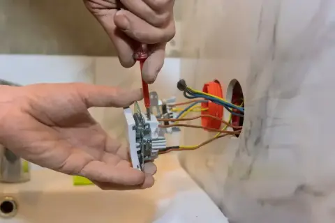 Elektryk montuje gniazdko, podłączając 3 kable. Widać jego dłonie i narzędzia.