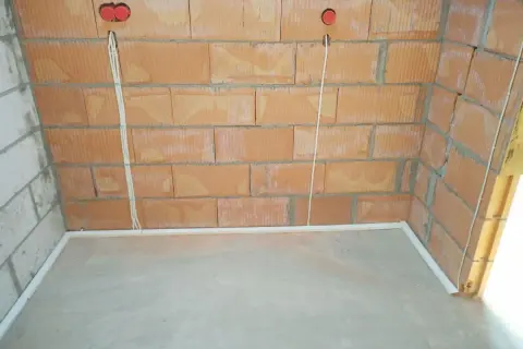 Widoczne przewody w posadzce bez peszla, biegnące od puszek instalacyjnych w ceglanej ścianie do podłogi.