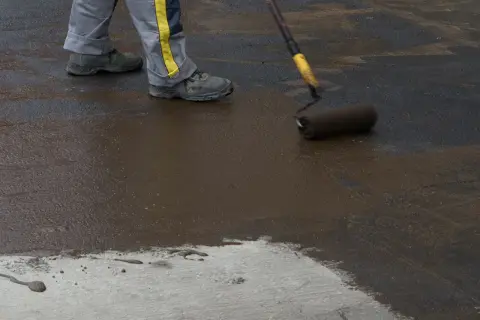 Pracownik maluje podłogę w piwnicy na gruncie ciemną masą. Widać jego nogi w roboczych spodniach i buty.