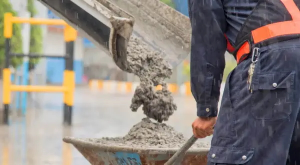 Robotnik w deszczu wylewa świeży beton do taczki. To właśnie jest beton – mieszanka cementu, kruszywa i wody.