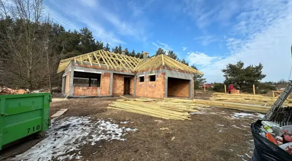Budowa domu z cegły, widoczny szkielet dachu, drewno budowlane, kontener na śmieci.