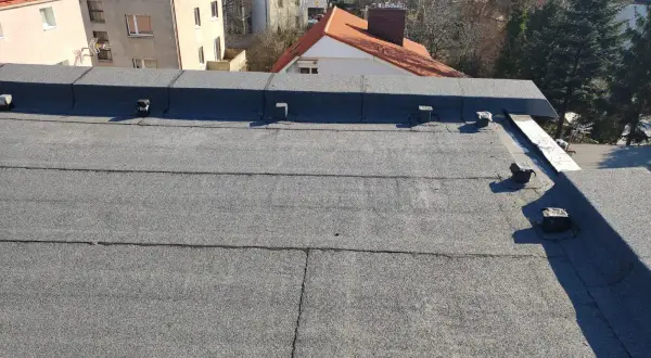 Praktyczny przykład, jak położyć papę termozgrzewalną na dachu. Widok na pokrycie dachowe i budynki w tle.