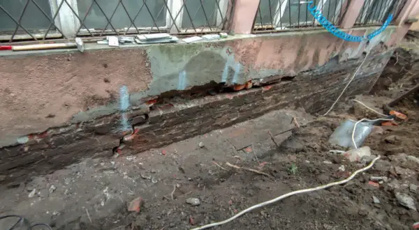 Widoczny pęknięty fundament z cegły, odsłonięty po wykopaniu ziemi. Widać ślady napraw i narzędzia.