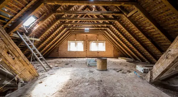 Poddasze 100m2 do adaptacji. Widok na drewnianą konstrukcję dachu, okna, drabiny i gruzy. Koszt adaptacji poddasza 100m2 może być wysoki.