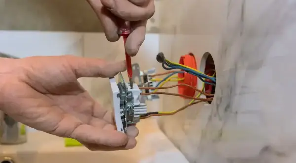 Elektryk montuje gniazdko, podłączając 3 kable. Widać jego dłonie i narzędzia.