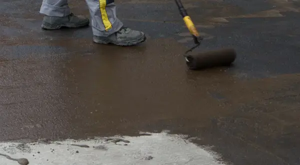 Pracownik maluje podłogę w piwnicy na gruncie ciemną masą. Widać jego nogi w roboczych spodniach i buty.