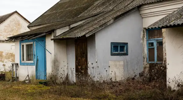 Stary budynek gospodarczy z niebieskimi drzwiami i oknami, porośnięty dziką roślinnością. Zastanawiasz się, jak zalegalizować budynek gospodarczy?