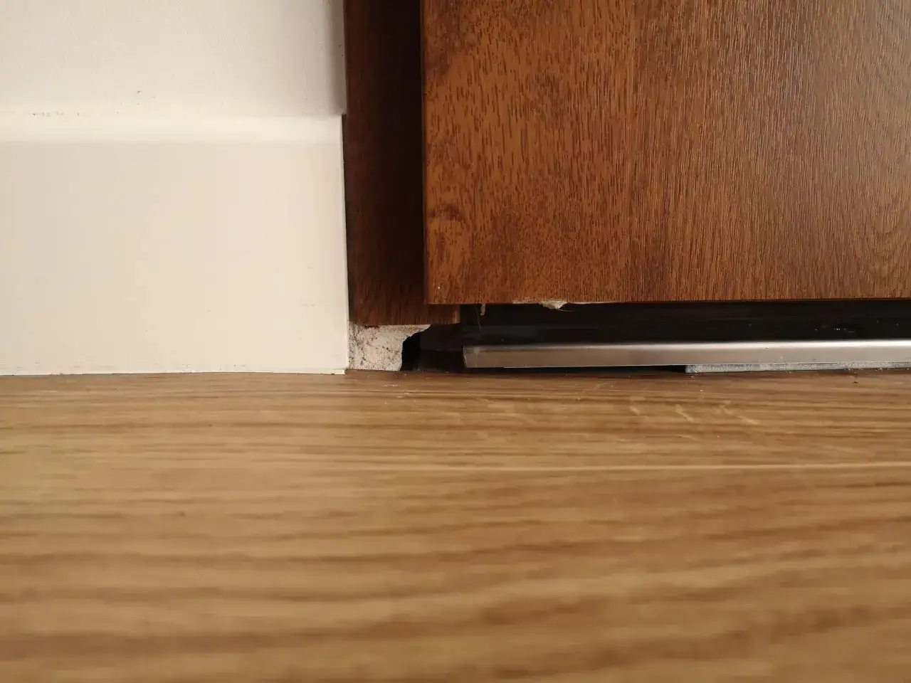 Widoczna szczelina między ościeżnicą a podłogą, z widocznym metalowym elementem i fragmentem drewna.