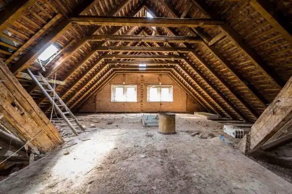 Poddasze 100m2 do adaptacji. Widok na drewnianą konstrukcję dachu, okna, drabiny i gruzy. Koszt adaptacji poddasza 100m2 może być wysoki.