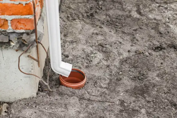 Biała rura spustowa odprowadza wodę do ceramicznej studzienki. Widać fragment ceglanego muru i betonowego cokołu.