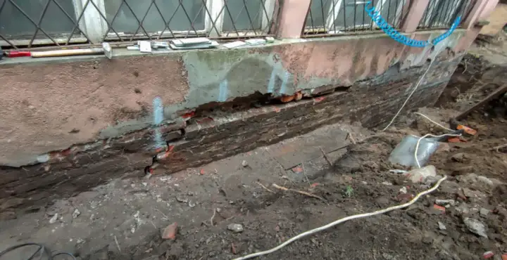 Widoczny pęknięty fundament z cegły, odsłonięty po wykopaniu ziemi. Widać ślady napraw i narzędzia.